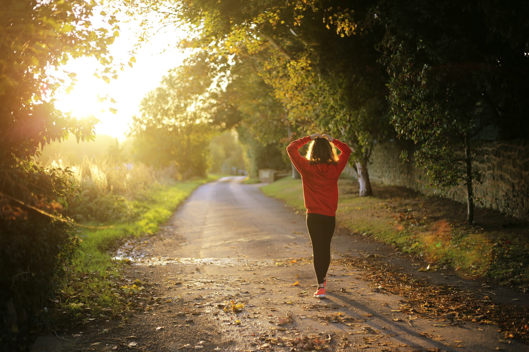 Woman in orange sweatshirt and gym clothes on country road with filtered sun through the trees